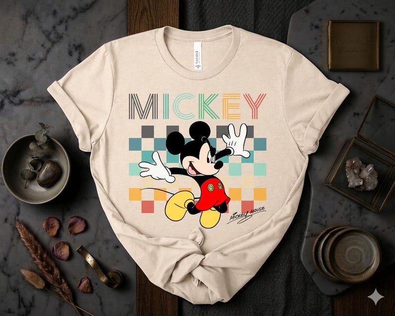 May include: Beige t-shirt featuring a colorful graphic of Mickey Mouse. The word "MICKEY" is printed across the top in various colors. Mickey Mouse is in a dynamic pose, with his arms outstretched. The shirt has a classic crew neck.