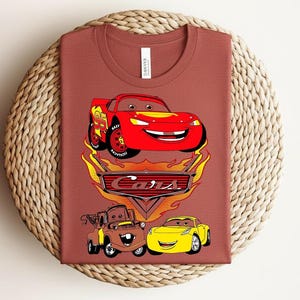 May include: A rust-colored t-shirt featuring a graphic of characters from the animated movie "Cars." The design includes a red race car, a brown tow truck, and a yellow sports car, with the word "Cars" in a stylized font.