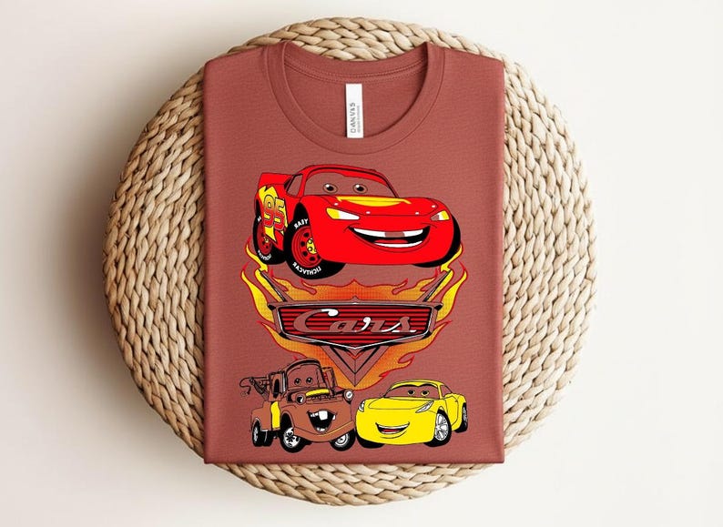 May include: A rust-colored t-shirt featuring a graphic of characters from the animated movie "Cars." The design includes a red race car, a brown tow truck, and a yellow sports car, with the word "Cars" in a stylized font.