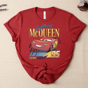 May include: A red t-shirt with a graphic of Lightning McQueen from the Cars film. The design includes the text "Lightning McQueen", a cartoon image of the car, and the number "95". The shirt also has the text "Think Fast".