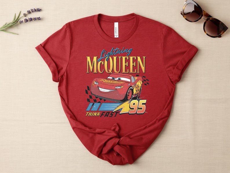 May include: A red t-shirt with a graphic of Lightning McQueen from the Cars film. The design includes the text "Lightning McQueen", a cartoon image of the car, and the number "95". The shirt also has the text "Think Fast".