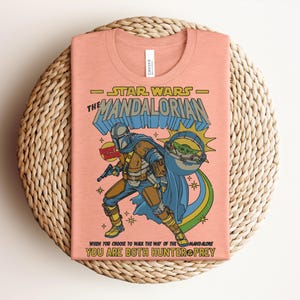 K&ouml;nnte beinhalten: Pfirsichfarbenes T-Shirt mit einer Grafik aus Star Wars: The Mandalorian. Das Design zeigt den Mandalorianer, Grogu und den Text "Star Wars The Mandalorian" und "When you choose to walk the way of the Mandalore, you are both hunter & prey."