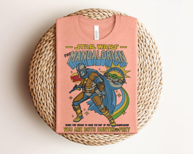 K&ouml;nnte beinhalten: Pfirsichfarbenes T-Shirt mit einer Grafik aus Star Wars: The Mandalorian. Das Design zeigt den Mandalorianer, Grogu und den Text "Star Wars The Mandalorian" und "When you choose to walk the way of the Mandalore, you are both hunter & prey."