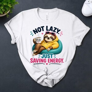 May include: White t-shirt with a cartoon sloth wearing sunglasses and a yellow hoodie, relaxing on a blue beanbag chair. The shirt has the text "NOT LAZY. JUST SAVING ENERGY." and a coffee cup that says "NOT FAST, NOT FURIOUS, JUST ME."