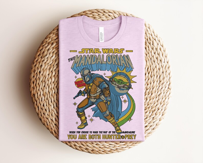 K&ouml;nnte beinhalten: Ein helllila T-Shirt mit einer Grafik von The Mandalorian aus Star Wars. Das Design zeigt den Mandalorianer in R&uuml;stung, Baby Yoda und den Text "STAR WARS THE MANDALORIAN". Das Shirt hat auch den Text "WHEN YOU CHOOSE TO WALK THE WAY OF THE MANDALORE YOU ARE BOTH HUNTER & PREY".