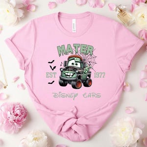 Może przedstawiać: Jasnor&oacute;żowa koszulka z halloweenowym motywem Złomka z filmu Auta Disneya. Projekt zawiera napisy "MATER", "EST 1977" i "Disney Cars". Obrazek przedstawia nietoperze i pajęczynę, tworząc upiorną estetykę.