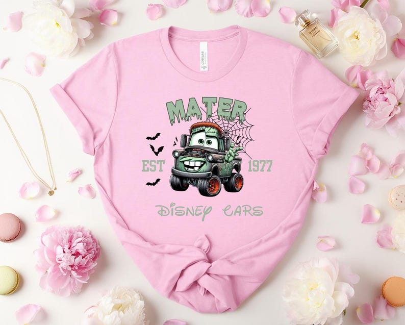 Może przedstawiać: Jasnor&oacute;żowa koszulka z halloweenowym motywem Złomka z filmu Auta Disneya. Projekt zawiera napisy "MATER", "EST 1977" i "Disney Cars". Obrazek przedstawia nietoperze i pajęczynę, tworząc upiorną estetykę.