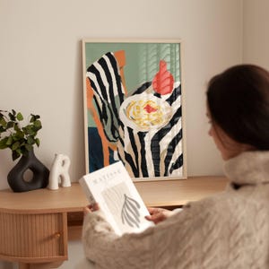 Puede incluir: Una impresión artística enmarcada que presenta una naturaleza muerta con un mantel a rayas de cebra, un plato de pasta y una botella roja. La obra de arte se exhibe en una pared sobre un escritorio de madera. Una persona sostiene un libro con la palabra "MATISSE" en la portada.