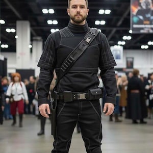 May include: A black costume with a high collar, shoulder strap, and utility belt. The outfit features white stitching and is paired with brown boots. The person is standing in a convention centre.