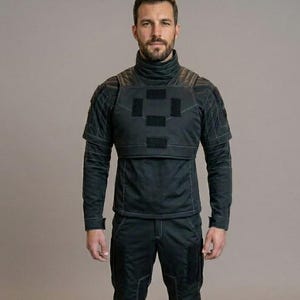 May include: A person wearing a black tactical outfit. The outfit includes a high-neck top with a quilted shoulder design and a square patch on the chest, long-sleeved shirt, and matching pants. The person is wearing black boots.