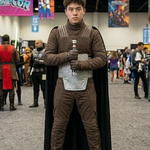May include: A person in a brown costume with a black cape, holding a lightsaber. The costume includes a high-collared jacket, trousers, and gloves. The person is standing in a convention hall with banners that say "Galaxy-Con" and "Kyber Kratters Booth".