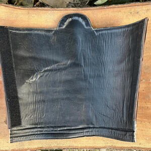 May include: A black leather item with a curved top and a rectangular shape. The item has a textured surface and a strip of Velcro on the left side. The item is laying on a wooden surface.