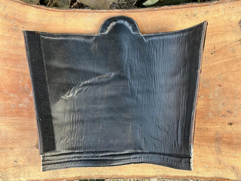 May include: A black leather item with a curved top and a rectangular shape. The item has a textured surface and a strip of Velcro on the left side. The item is laying on a wooden surface.