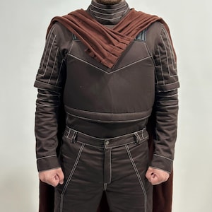 May include: A brown costume with white stitching, featuring a high-collared top, a cape, and matching pants. The costume has a padded appearance and is designed with a geometric pattern. The cape is a darker shade of brown.