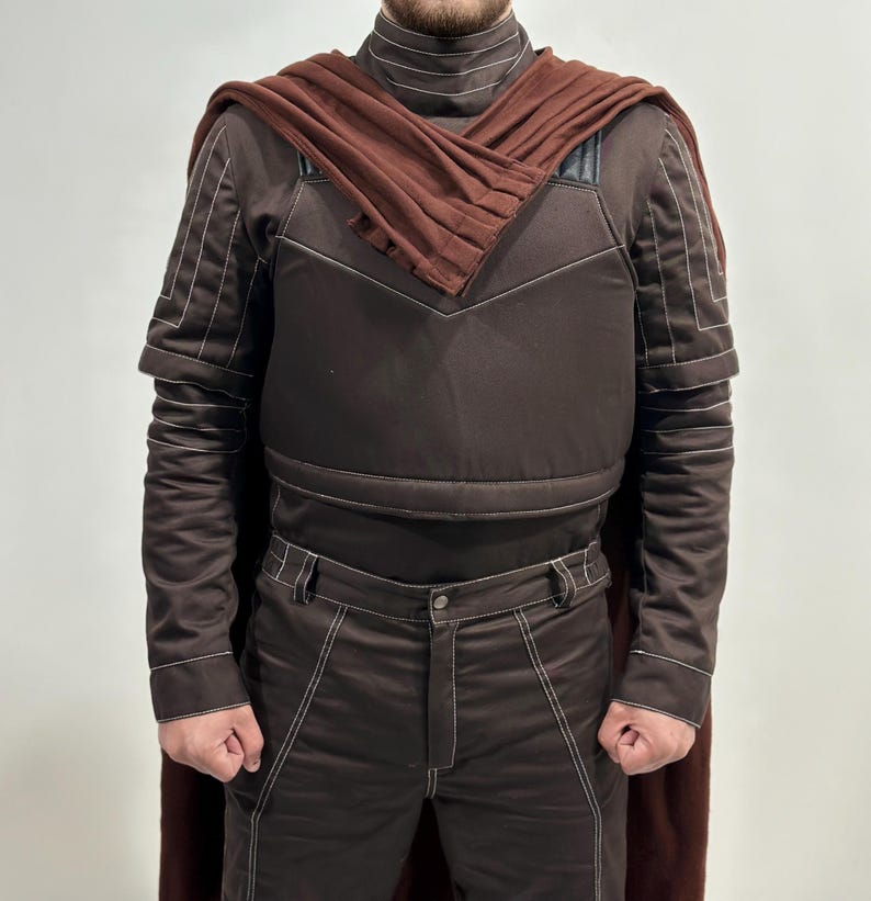 May include: A brown costume with white stitching, featuring a high-collared top, a cape, and matching pants. The costume has a padded appearance and is designed with a geometric pattern. The cape is a darker shade of brown.