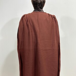 May include: A dark reddish-brown cape with a high collar, viewed from the back. The cape is made of a thick fabric and drapes down to the ankles. The collar is a lighter brown with a striped pattern.