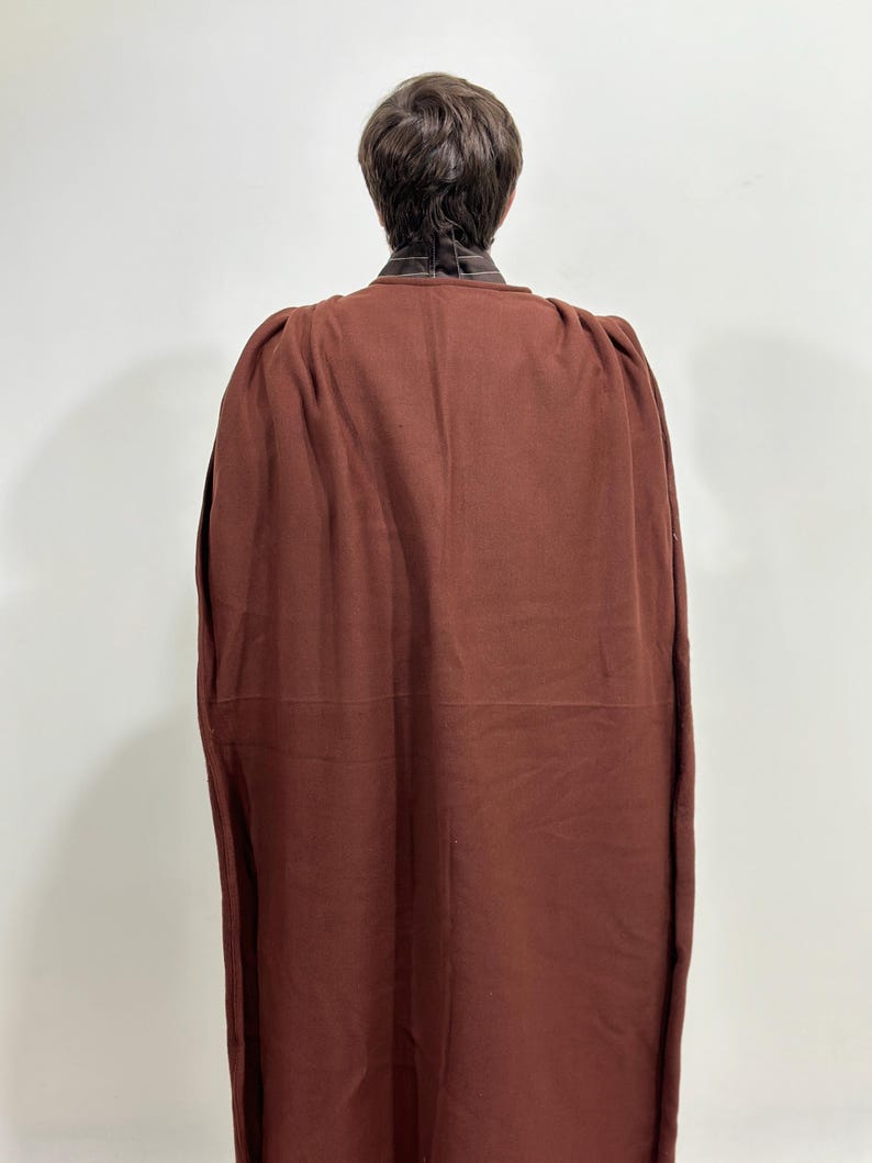 May include: A dark reddish-brown cape with a high collar, viewed from the back. The cape is made of a thick fabric and drapes down to the ankles. The collar is a lighter brown with a striped pattern.