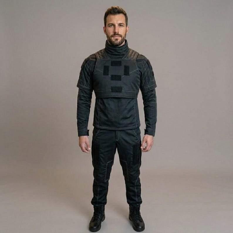 May include: A person wearing a black tactical outfit. The outfit includes a high-neck top with a quilted shoulder design and a square patch on the chest, long-sleeved shirt, and matching pants. The person is wearing black boots.