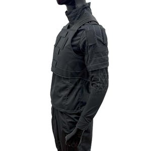 May include: Black tactical vest with multiple rectangular pockets and a high collar. The vest is worn over a long-sleeved shirt and pants. White stitching accents the seams and geometric patterns on the sleeves.