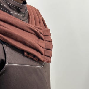 May include: A brown garment with white stitching details. The garment features a high collar and a pleated, rust-colored shoulder wrap. The fabric appears to be a soft, textured material. The overall style suggests a modern, minimalist design.