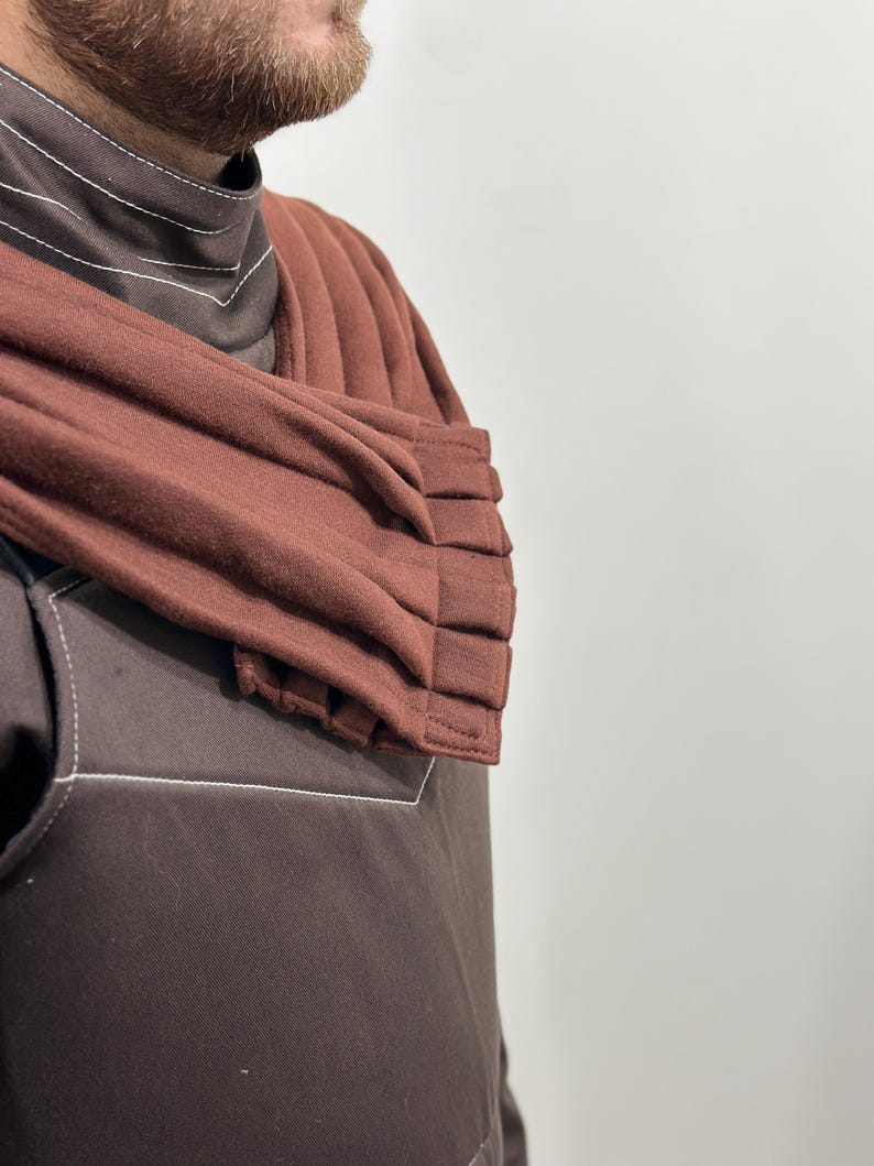 May include: A brown garment with white stitching details. The garment features a high collar and a pleated, rust-colored shoulder wrap. The fabric appears to be a soft, textured material. The overall style suggests a modern, minimalist design.