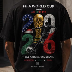 May include: A black t-shirt featuring a FIFA World Cup 2026 graphic on the back. The design includes the text FIFA WORLD CUP 2026, the trophy, a soccer player, a soccer ball, and the flags of the United States, Mexico, and Canada.