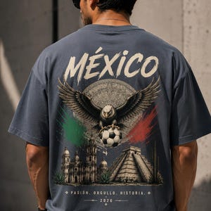 May include: A slate blue t-shirt featuring a large back graphic with the word M&Eacute;XICO in white. The design includes a bald eagle holding a soccer ball, a circular calendar stone, a cathedral, a pyramid, and green and red brushstroke accents.