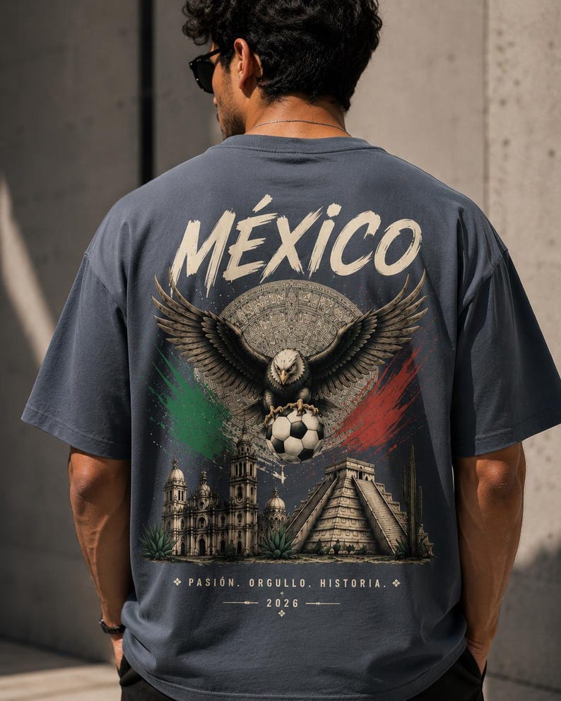 May include: A slate blue t-shirt featuring a large back graphic with the word M&Eacute;XICO in white. The design includes a bald eagle holding a soccer ball, a circular calendar stone, a cathedral, a pyramid, and green and red brushstroke accents.