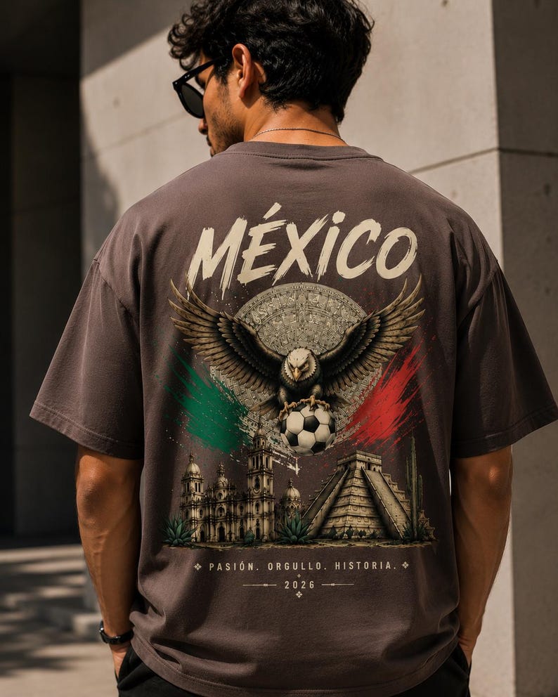 May include: A charcoal gray t-shirt featuring a back graphic with the word M&Eacute;XICO in large letters, an eagle clutching a soccer ball, a circular calendar stone, green and red brushstrokes, architectural landmarks, and the text PASI&Oacute;N. ORGULLO. HISTORIA. 2026.