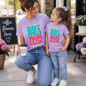 以下が含まれることがあります： 「HOT MESS MOM CLUB」と「HOT MESS MINI CLUB」の文字がカラフルにプリントされた、お揃いのラベンダー色のTシャツ。女性は「mom fuel」と書かれたコーヒーカップを持っています。どちらも青いジーンズと白いスニーカーを着用しています。
