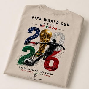 May include: A beige t-shirt featuring a FIFA World Cup 2026 graphic. The design includes the text, the flags of the United States, Mexico, and Canada, a gold trophy, a soccer player, a soccer ball, and the text Three Nations. One Dream.