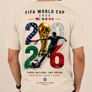 May include: A beige t-shirt featuring a FIFA World Cup 2026 graphic on the back. The design includes the flags of the USA, Mexico, and Canada, a gold trophy, a soccer player silhouette, a soccer ball, and the text Three Nations. One Dream.