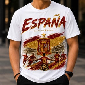 May include: A white short-sleeved t-shirt featuring a graphic print with the text ESPAÑA and LA FURIA ROJA. The design includes a gold star, the Spanish coat of arms, a soccer stadium background, and soccer players in red jerseys with numbers.
