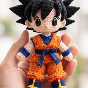 Puede incluir: Muñeca de ganchillo hecha a mano de un personaje con pelo negro puntiagudo, vistiendo un atuendo naranja con detalles azules. La muñeca se sostiene en una mano, mostrando la detallada artesanía y los colores vibrantes. La muñeca tiene ojos grandes y expresivos.