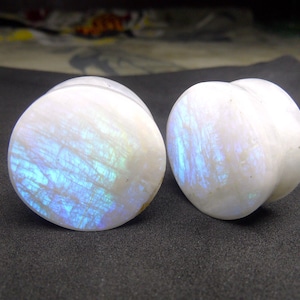 Könnte beinhalten: Zwei weiße, irisierende Mondstein-Ohrstecker. Einer ist rund, der andere hat die Form eines Pfirsichs. Die Ohrstecker haben einen perlmuttartigen Glanz mit blauen und grünen Reflexen. Der Hintergrund ist ein dunkelgrauer Stoff.