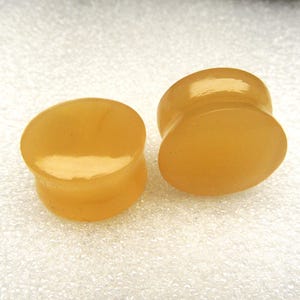 May include: A pair of smooth, round ear plugs with a polished finish and a translucent, warm honey-yellow color. These circular body jewelry pieces feature a concave center design with flared edges, showcasing a solid, uniform appearance throughout.