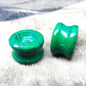 Puede incluir: Dos tapones de piedra de malaquita verde. Uno es cilíndrico con base y parte superior ensanchadas, el otro es un túnel curvo. Los tapones tienen un acabado pulido y brillante y un distintivo patrón de bandas verde claro y oscuro.