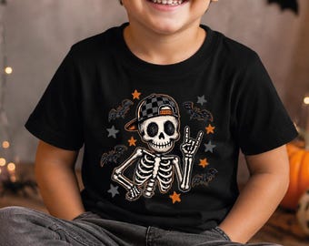 May include: A black short-sleeve t-shirt featuring a graphic of a skeleton wearing a checkered baseball cap and making a hand gesture, surrounded by small orange and gray stars and bats, paired with gray denim pants and black and white sneakers.