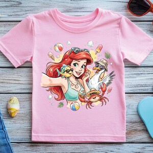Puede incluir: Camiseta rosa claro con una ilustraci&oacute;n de dibujos animados de un personaje pelirrojo tom&aacute;ndose una selfie con un pez, un cangrejo y una gaviota. La imagen est&aacute; rodeada de accesorios de playa, incluyendo gafas de sol y chanclas.