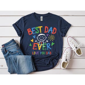 May include: Navy blue t-shirt with the text "BEST DAD EVER LOVE YOU DAD" in colorful, playful lettering. The design includes a stick figure, hearts, and a sun. The shirt is paired with blue jeans and white sneakers.