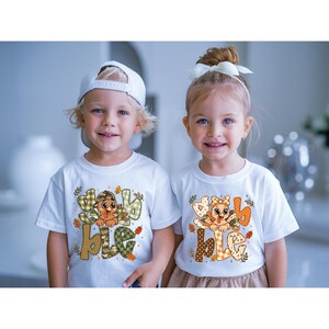May include: Two white t-shirts featuring a graphic of a cartoon turkey and the text "gobble". The left shirt shows a turkey in a camouflage cap with green plaid lettering. The right shirt shows a turkey with a bow and orange patterned lettering.