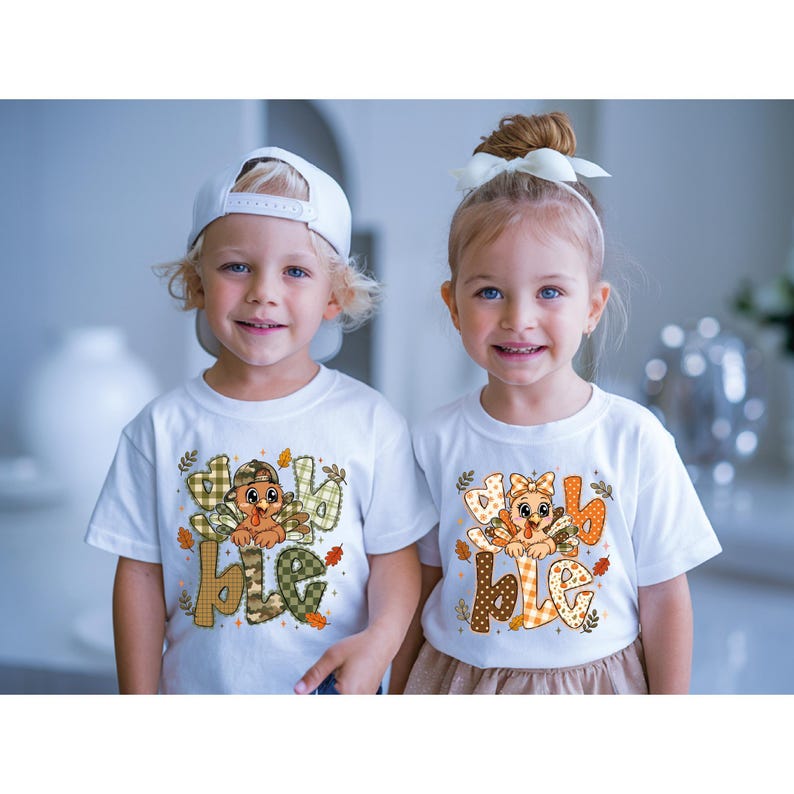 May include: Two white t-shirts featuring a graphic of a cartoon turkey and the text "gobble". The left shirt shows a turkey in a camouflage cap with green plaid lettering. The right shirt shows a turkey with a bow and orange patterned lettering.