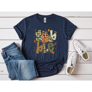 May include: A navy blue short-sleeve t-shirt featuring a graphic of a cartoon turkey wearing a camouflage baseball cap. The turkey is positioned behind the word "dabble" written in large, patterned letters with plaid and camouflage designs.