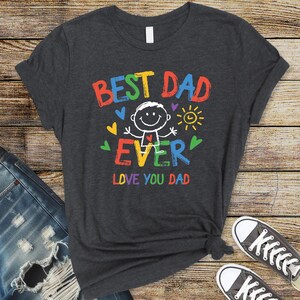 May include: Dark gray t-shirt with the words "BEST DAD EVER" in colorful block letters. A stick figure with a smiling face is in the center, with hearts and a sun graphic. The text "LOVE YOU DAD" is below the graphic.