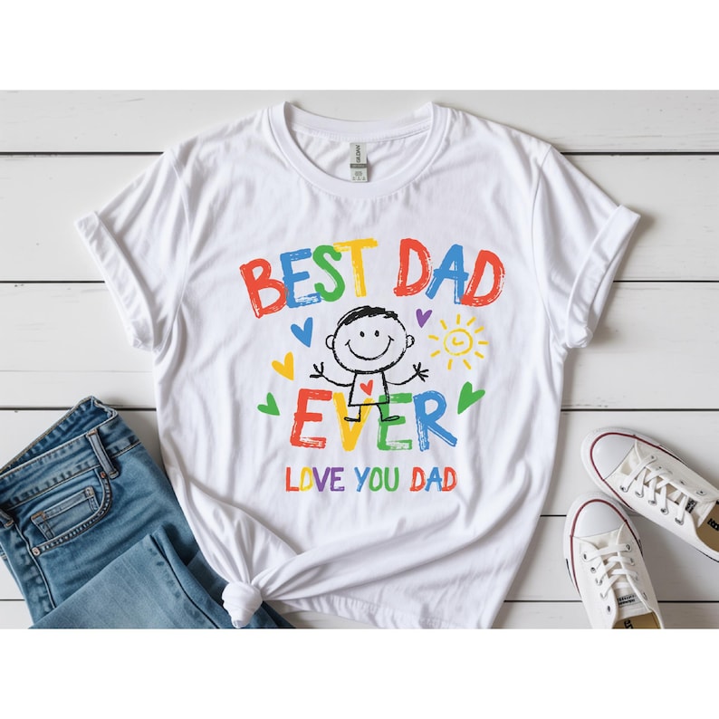 May include: White t-shirt with the text "BEST DAD EVER" in colorful letters, along with a stick figure illustration and the words "LOVE YOU DAD." The design includes hearts and a sun. The shirt is paired with blue jeans and white sneakers.