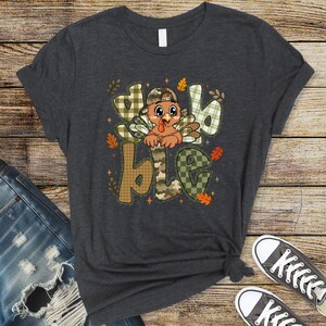 May include: A dark charcoal gray short-sleeve t-shirt featuring a graphic of a cartoon turkey wearing a camouflage baseball cap. The turkey is centered within the large, block-lettered text "gobble" which is filled with green and tan plaid patterns.