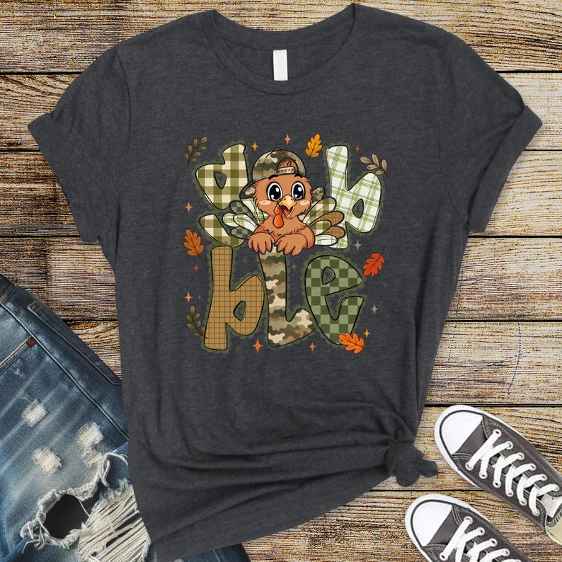 May include: A dark charcoal gray short-sleeve t-shirt featuring a graphic of a cartoon turkey wearing a camouflage baseball cap. The turkey is centered within the large, block-lettered text "gobble" which is filled with green and tan plaid patterns.