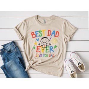 May include: A tan t-shirt with the words "BEST DAD EVER LOVE YOU DAD" in colorful letters. A cartoon figure is in the center of the design, with hearts and a sun. The shirt is paired with blue jeans and white sneakers.
