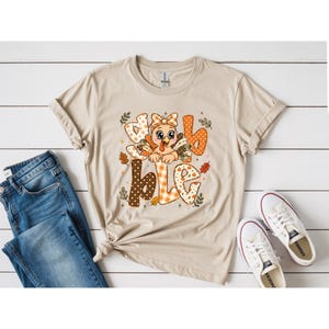 May include: A beige short-sleeved t-shirt featuring a graphic of a cartoon turkey with a bow, surrounded by the text "gobble" in various orange, brown, and white patterns. The design includes small autumn leaves and stars in a fall color palette.