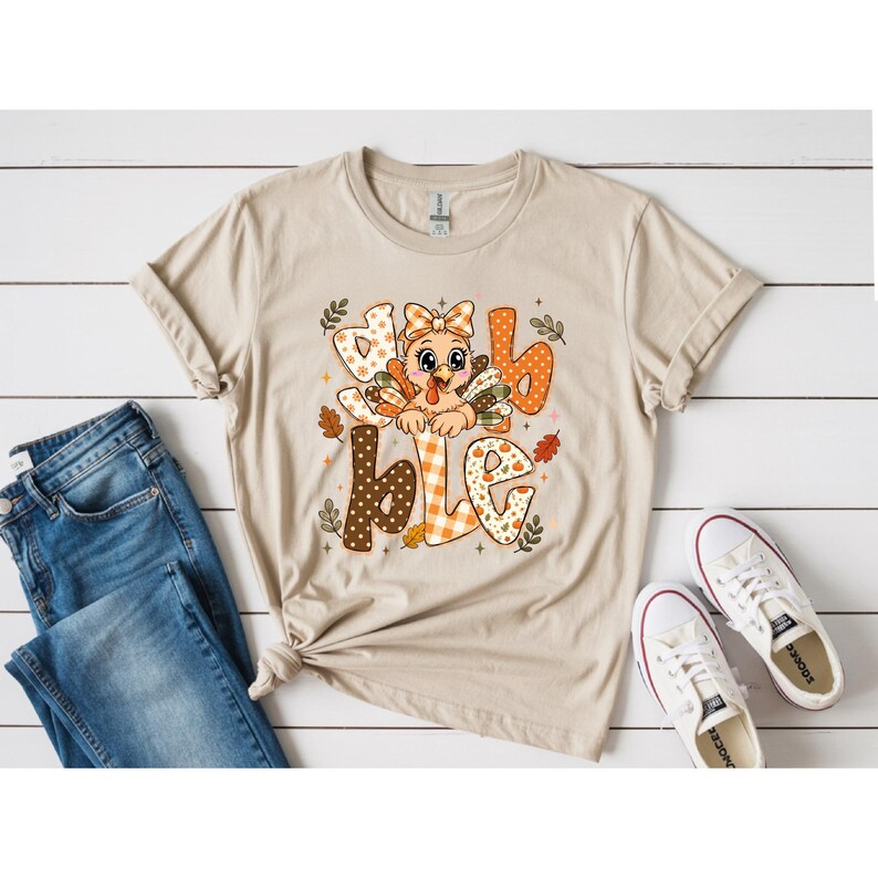 May include: A beige short-sleeved t-shirt featuring a graphic of a cartoon turkey with a bow, surrounded by the text "gobble" in various orange, brown, and white patterns. The design includes small autumn leaves and stars in a fall color palette.